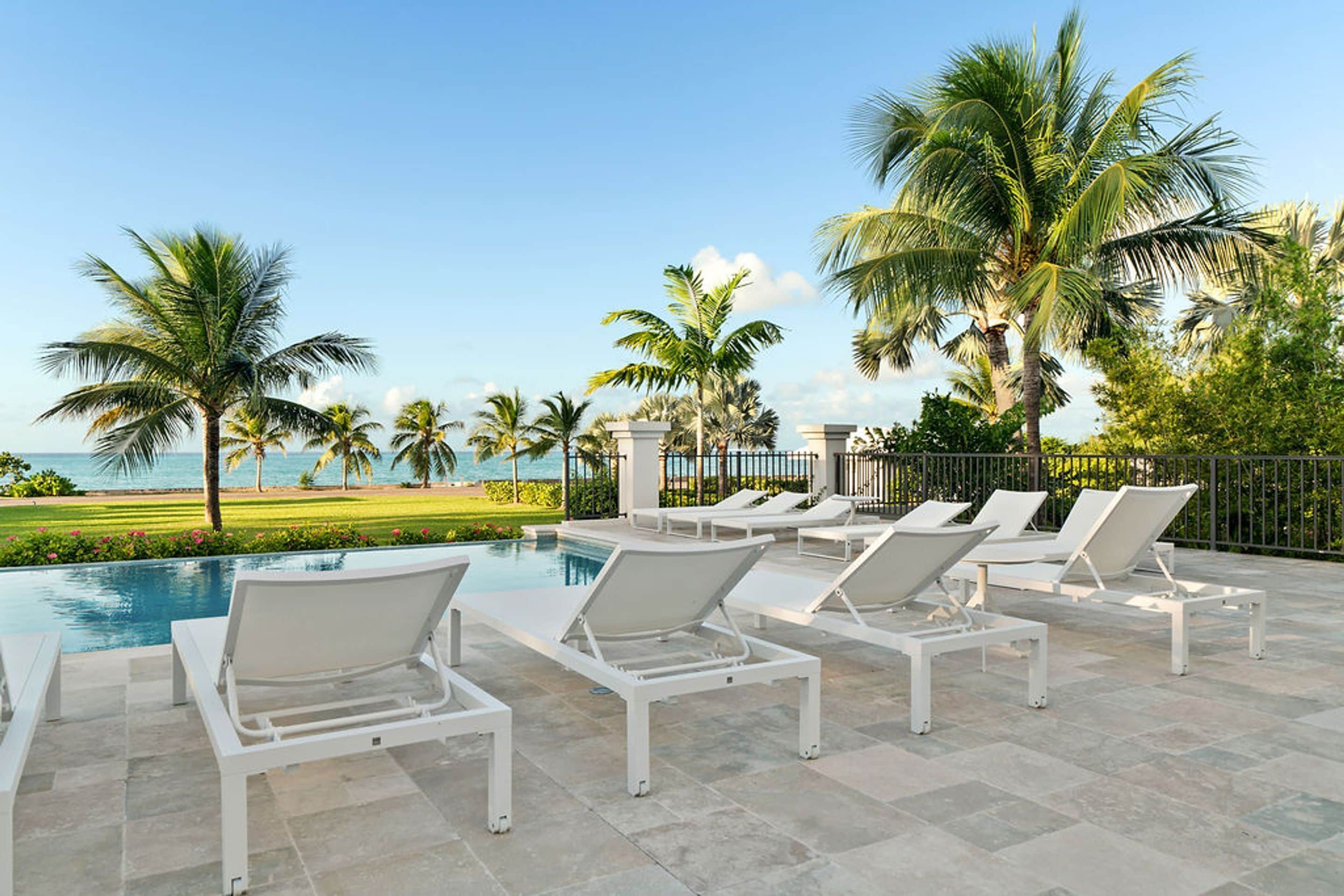 A poolside area with white lounge chairs, a pool, and a view of the ocean and palm trees.