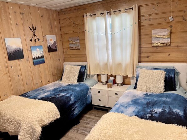 Una habitación acogedora con paredes de madera, dos camas con ropa de cama azul a juego, almohadas blancas mullidas y un ventanal con cortinas traslúcidas.