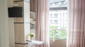 Un dormitorio con cama, armario, una mesita con una planta y ventana con cortinas.