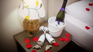 Una cama con pétalos de rosa, una botella de champán, una lámpara y bombones sobre una mesita de noche.