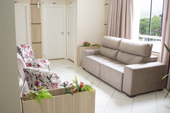 A living room with a beige sofa, a floral armchair, and a wooden side table with flowers. There are two doors in the background.
