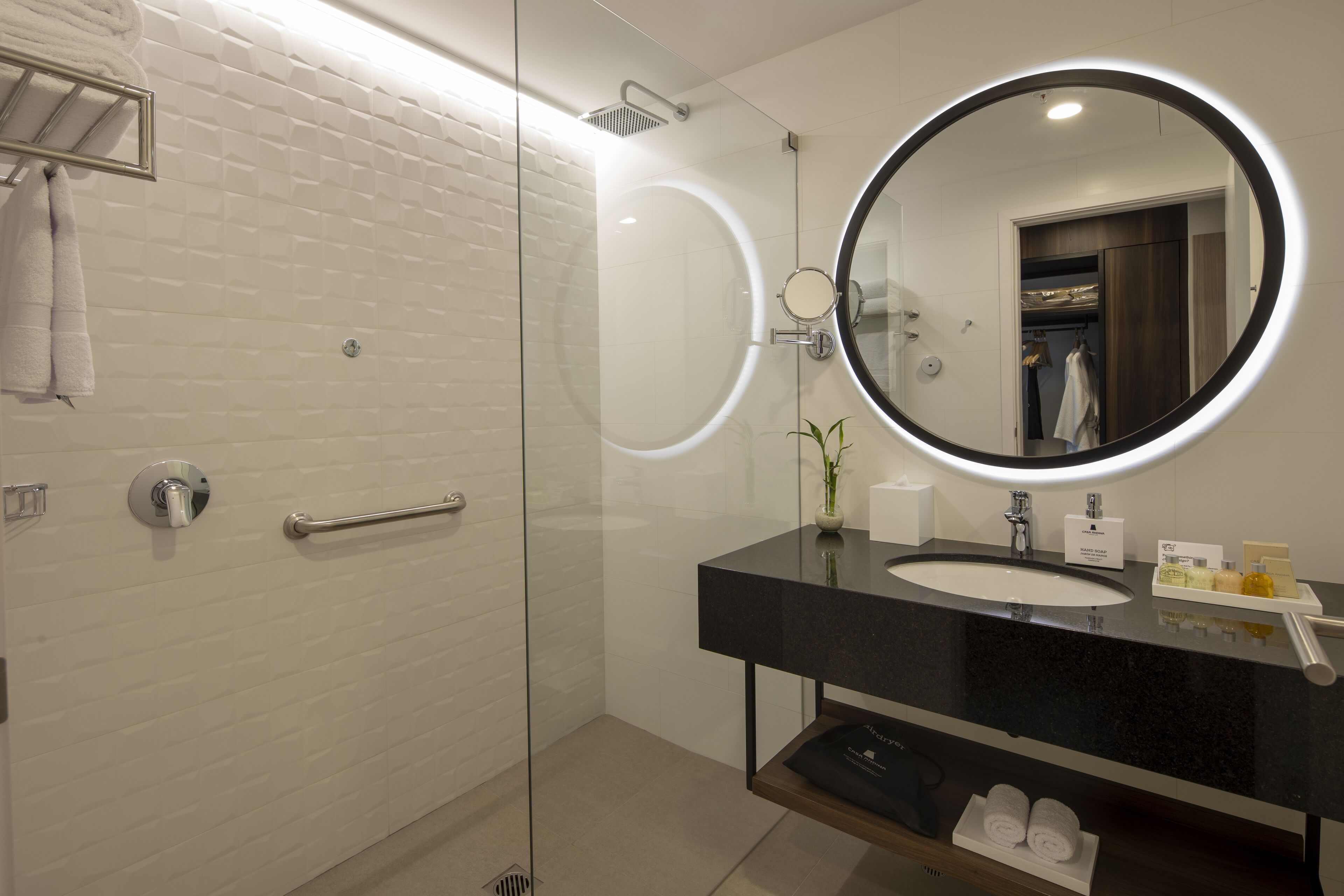 Un baño moderno con ducha de vidrio, un espejo redondo con marco negro y una encimera negra con lavabo y artículos de aseo.