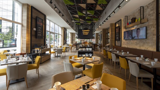 Een modern café met geel en grijs zitmeubilair, een centraal bargedeelte en grote ramen die voor natuurlijk licht zorgen.