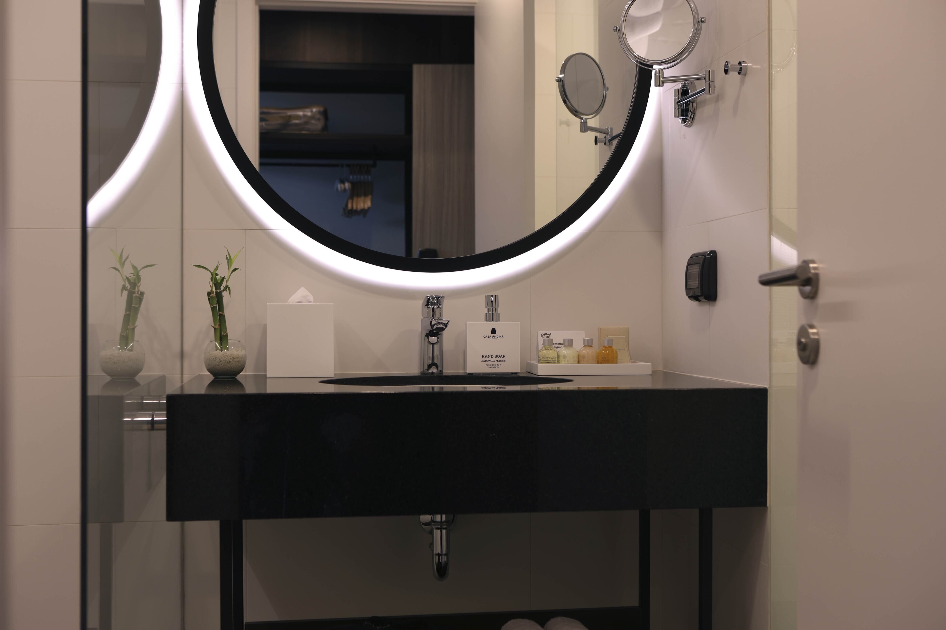 Une salle de bain moderne avec un lavabo noir, un miroir rond avec un éclairage intégré et deux plantes en pot.