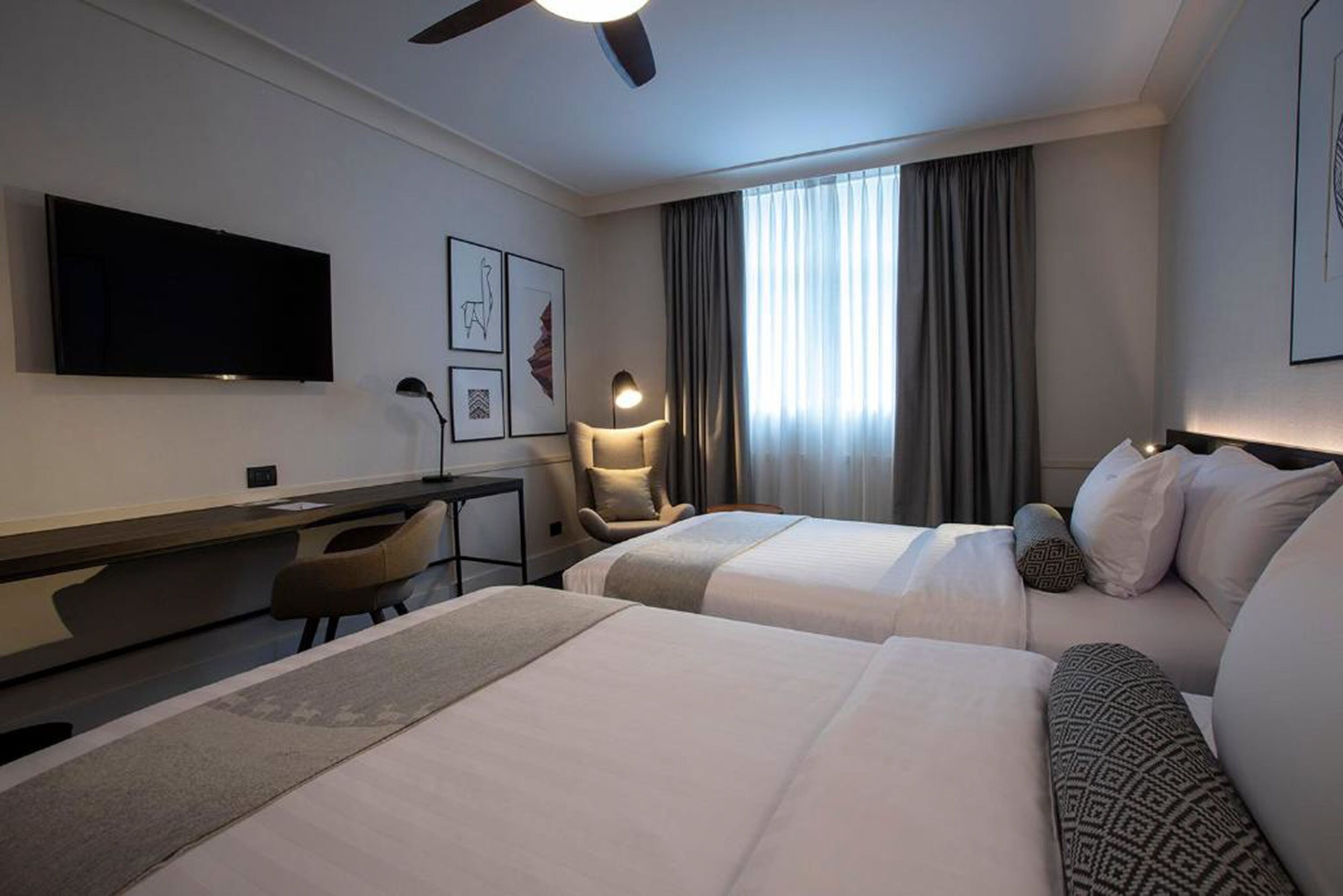 Habitación de hotel con dos camas, un escritorio, una silla, un televisor y un ventanal con cortinas.