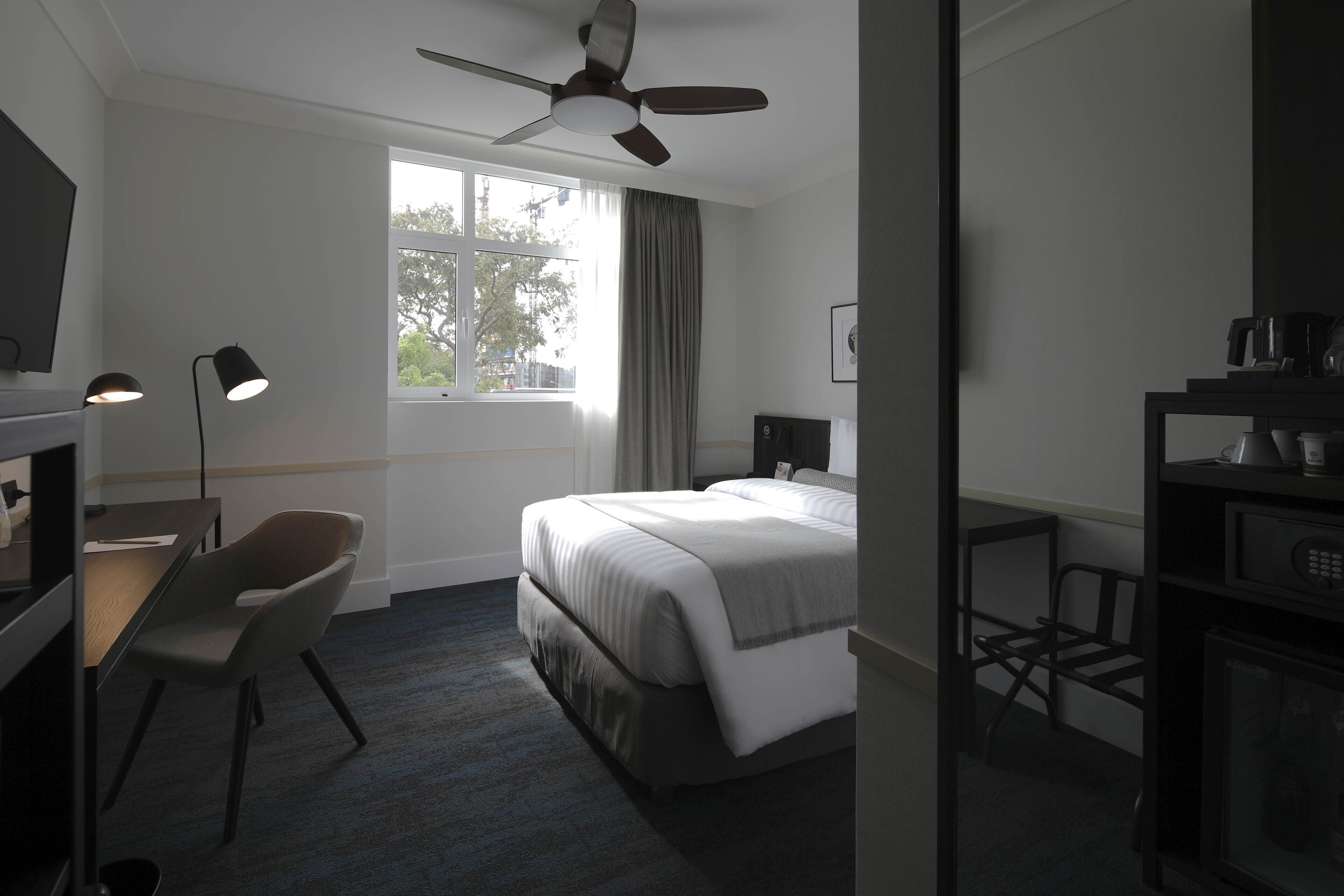 A hotel room with a bed, desk, chair, ceiling fan, and a window with curtains.