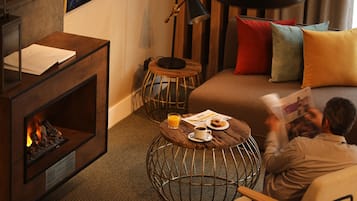 Una habitación acogedora con chimenea, una mesa de centro redonda con una bebida y un plato de comida, un sofá cómodo con cojines de colores, y una persona leyendo un libro.
