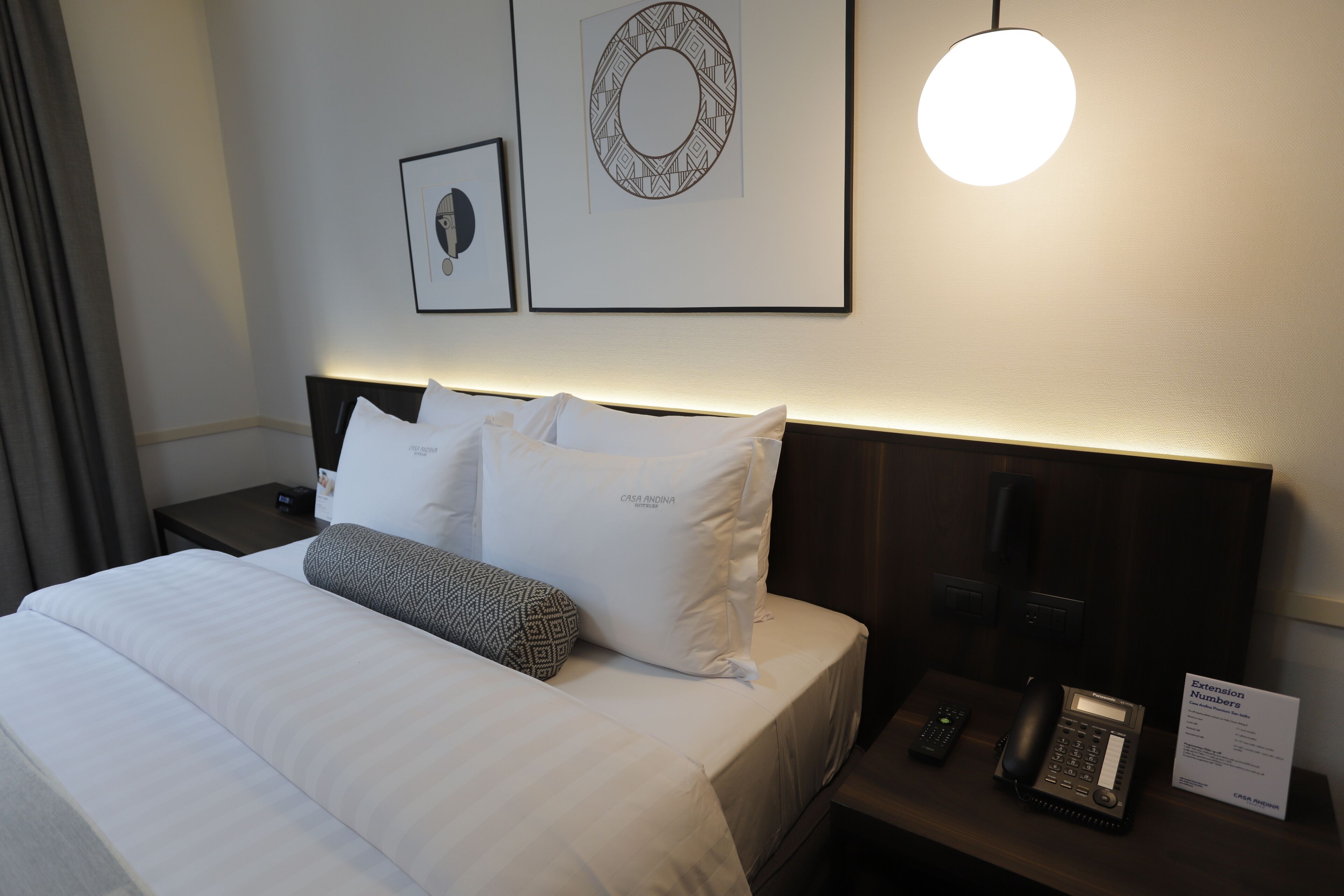 A hotel room with a neatly made bed, a bedside table with a phone and a sign, and wall art.