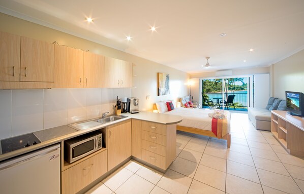 A modern kitchen with a built-in microwave, a sink, and wooden cabinets. There is a bed with a red pillow and a view of the ocean through large windows.