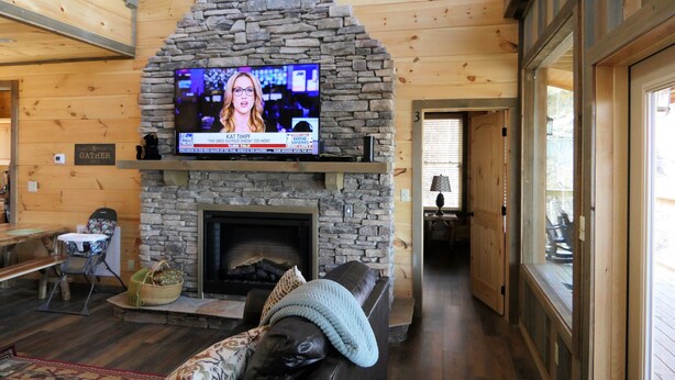 A cozy cabin interior with a stone fireplace, a television mounted above it, and a wooden dining table with chairs.