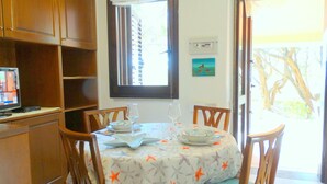 A dining area with a table set for two, wooden chairs, a television on a shelf, and a window with blinds. - Villa dell'Arte, on the beach of Porto Sa Ruxi, Villasimius (Porto Sa Ruxi)