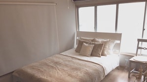 Um quarto com cama, janela, cadeira e uma pequena mesa.