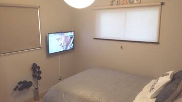 A bedroom with a bed, a television mounted on the wall, a window with a 'PEACE' sign, and a small plant on the floor.