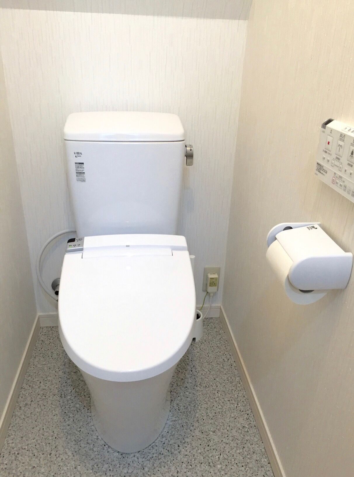 A small, white toilet with a control panel on the wall, a paper towel dispenser, and a toilet paper holder.