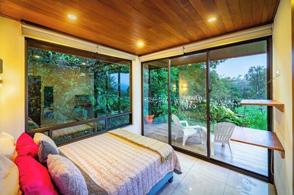 Un dormitorio con una cama grande, vistas a exuberante vegetación a través de ventanales del suelo al techo y un balcón con mobiliario de exterior.
