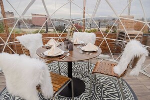 A round table set for four with white napkins, plates, and cutlery. There are four white furry chairs around the table. The table is on a patterned rug, and the setting is outdoors with a view of buildings in the background.
