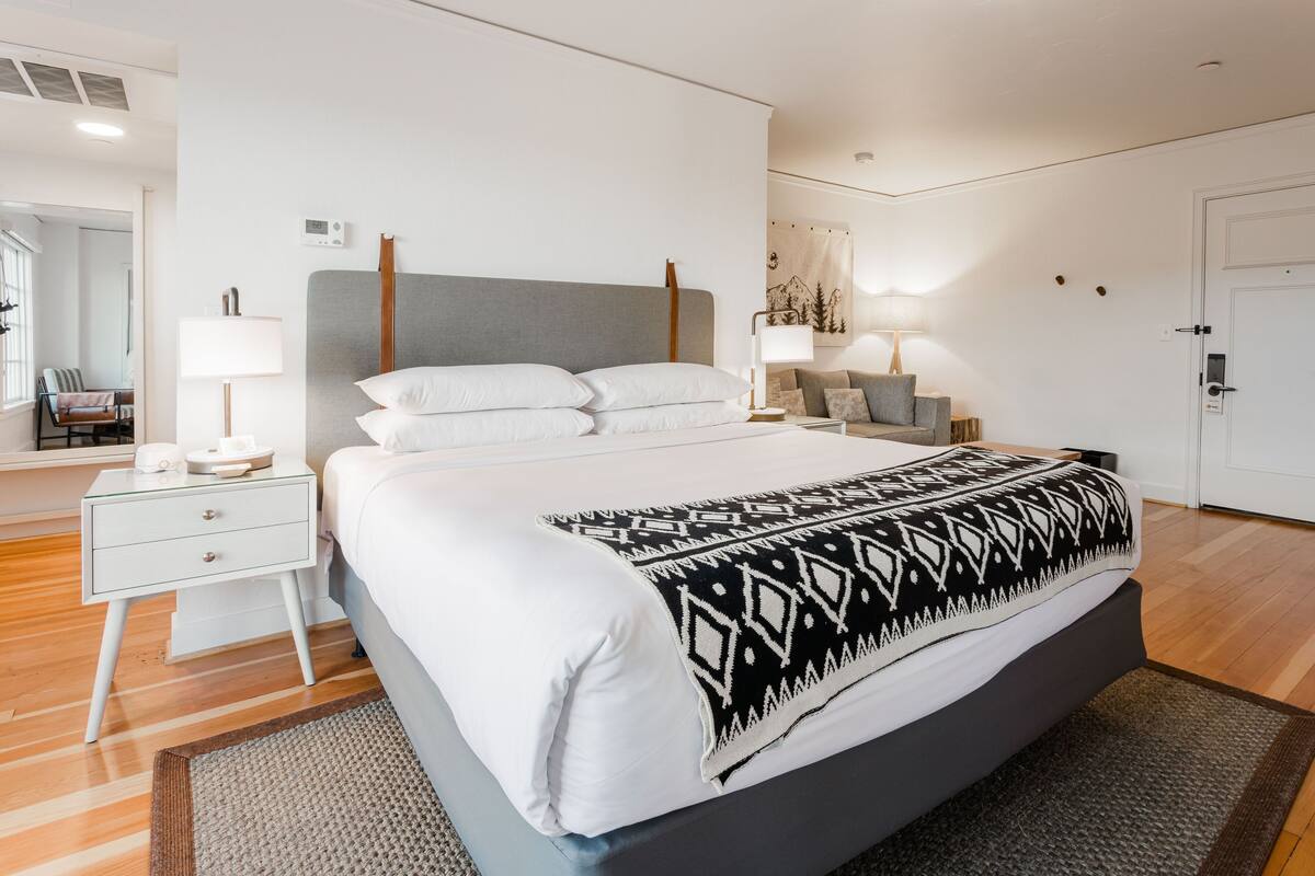 A modern bedroom with a large bed, bedside tables, a nightstand, a lamp, and a wall-mounted thermostat.