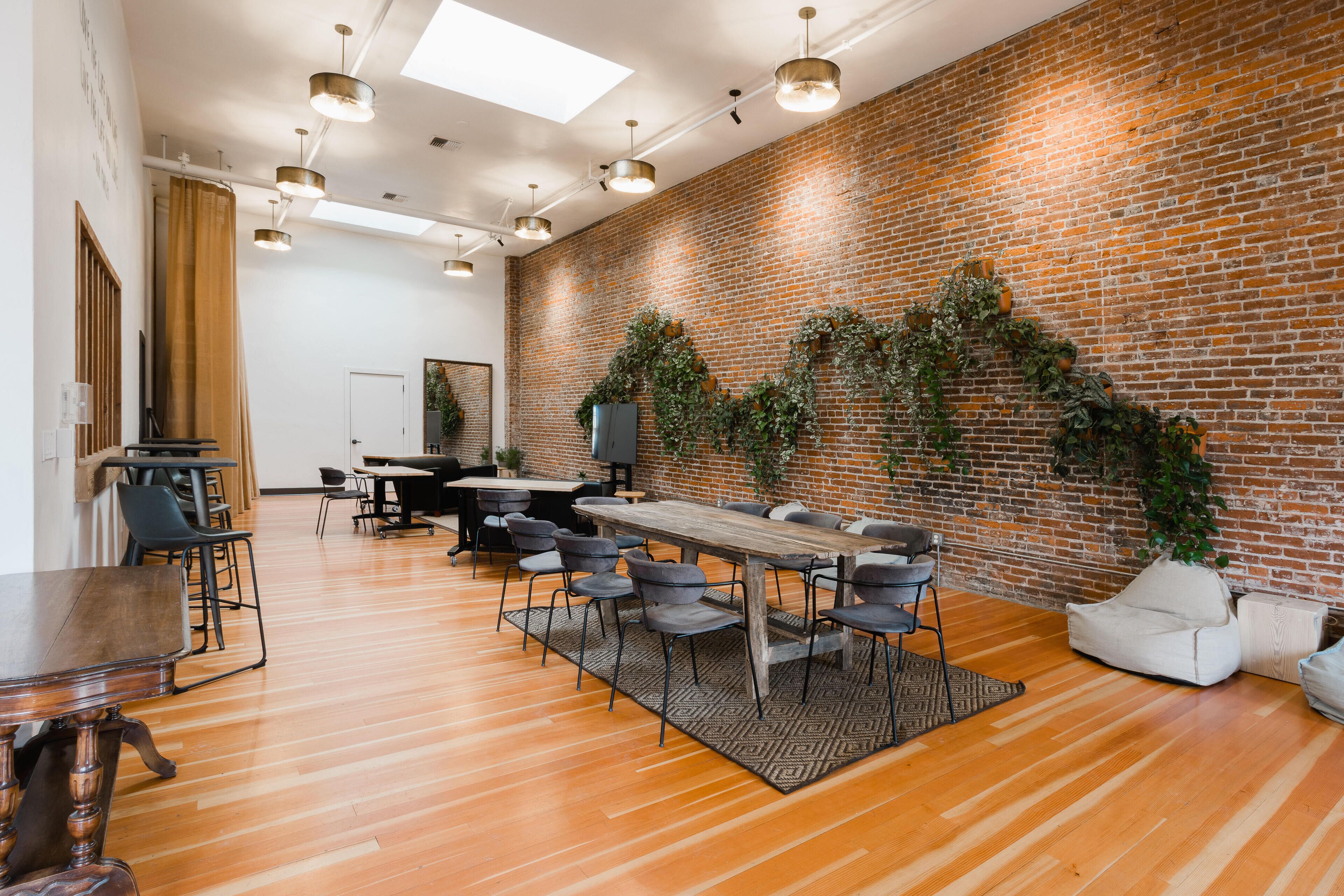 A modern meeting room with a brick wall, wooden tables, and chairs, featuring a large window and ceiling lights.