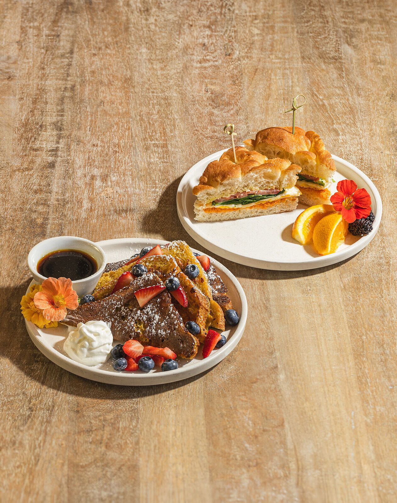 Two plates of food, one with pancakes and the other with a sandwich, both on a wooden table.