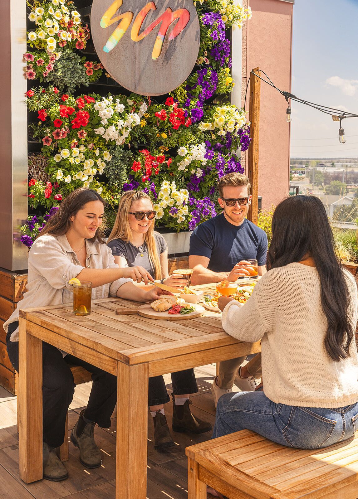 A group of people dining at an outdoor wooden table with a flower wall behind them.