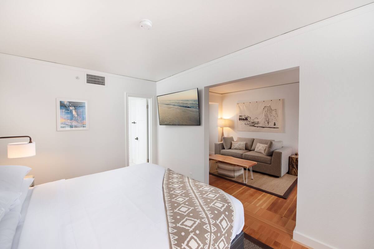 A modern hotel room with a large bed, a sofa, and a dining table. There are wall art pieces and a ceiling vent.