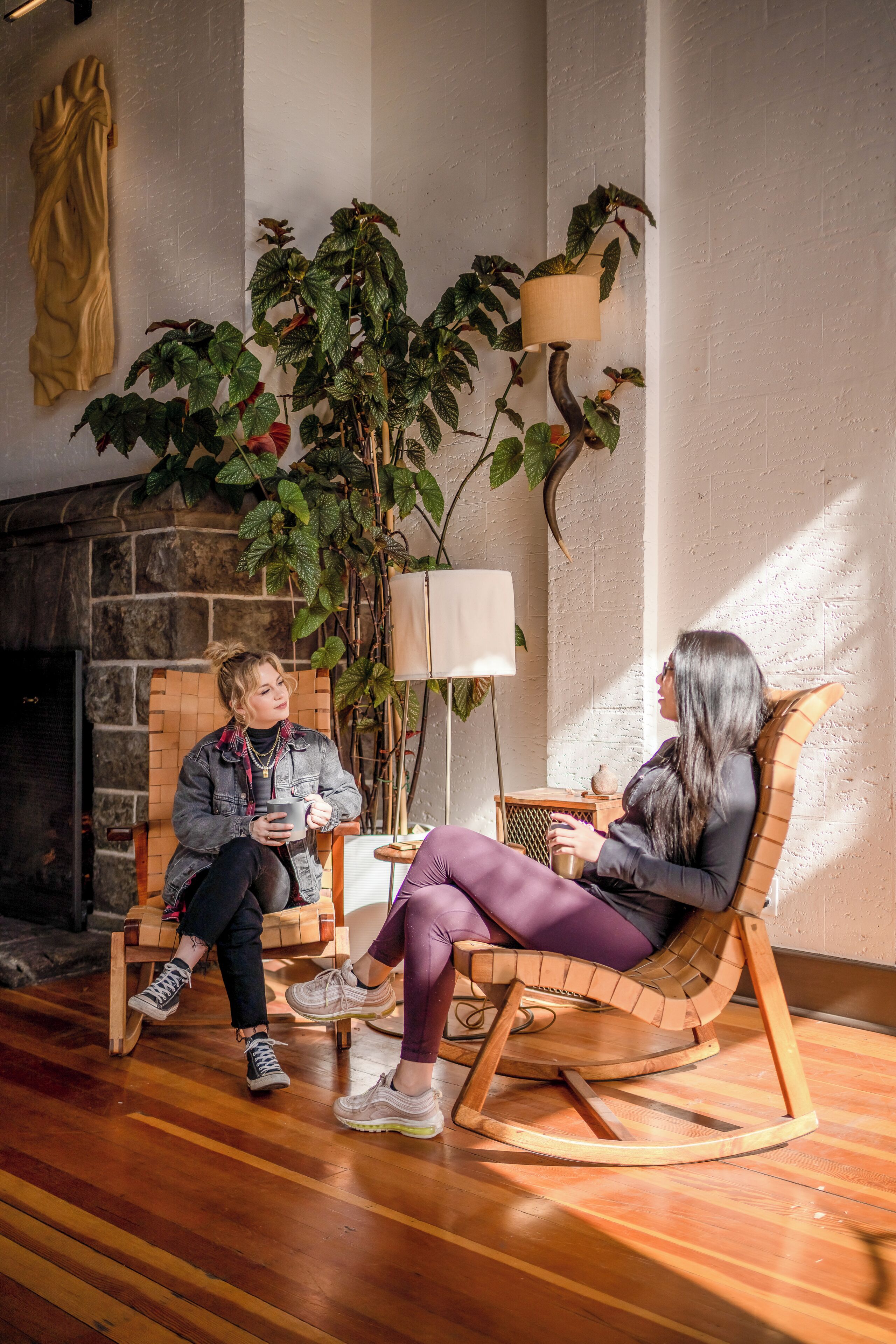 Two people seated on rocking chairs in a room with a fireplace, wooden flooring, and potted plants.
