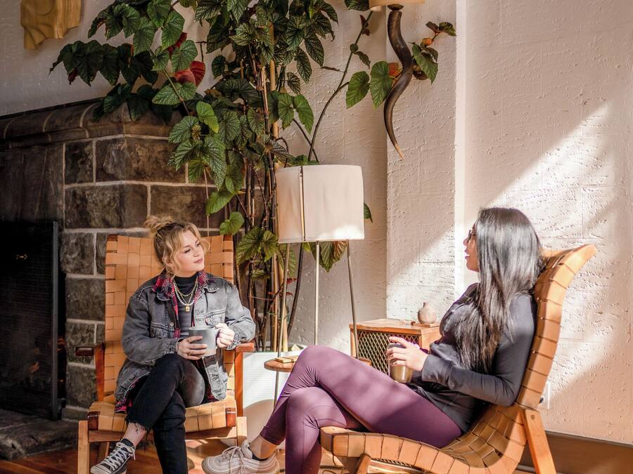 Two people seated on rocking chairs in a room with a fireplace, wooden flooring, and potted plants.