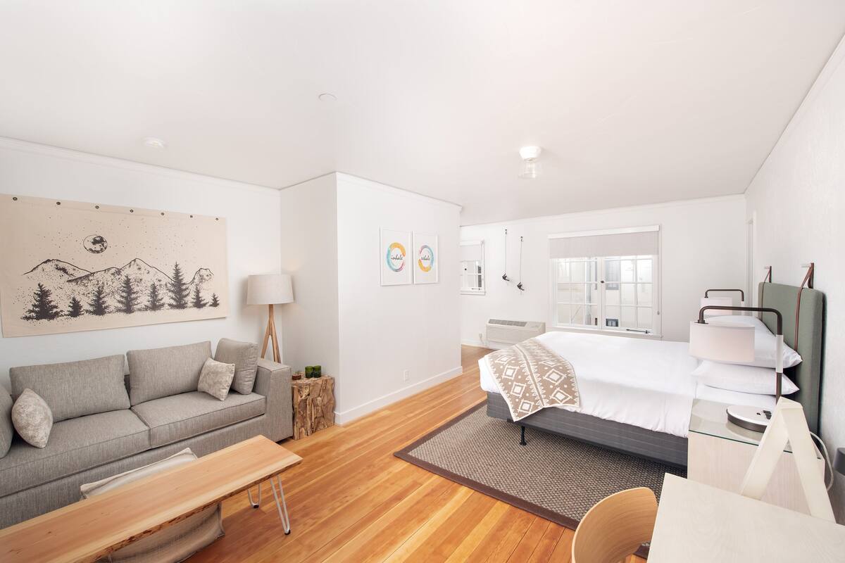 A modern hotel room with a grey sofa, a wooden coffee table, a bed with a patterned bedspread, and a desk with a chair.