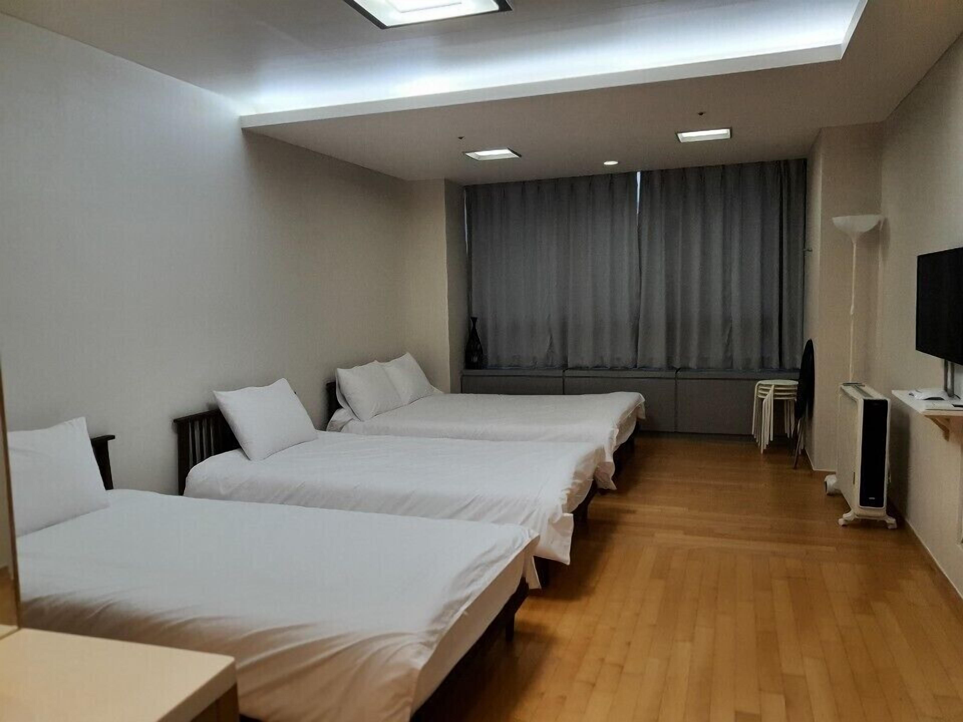 A hotel room with three single beds, a flat-screen TV, a large window with curtains, and a wooden floor.
