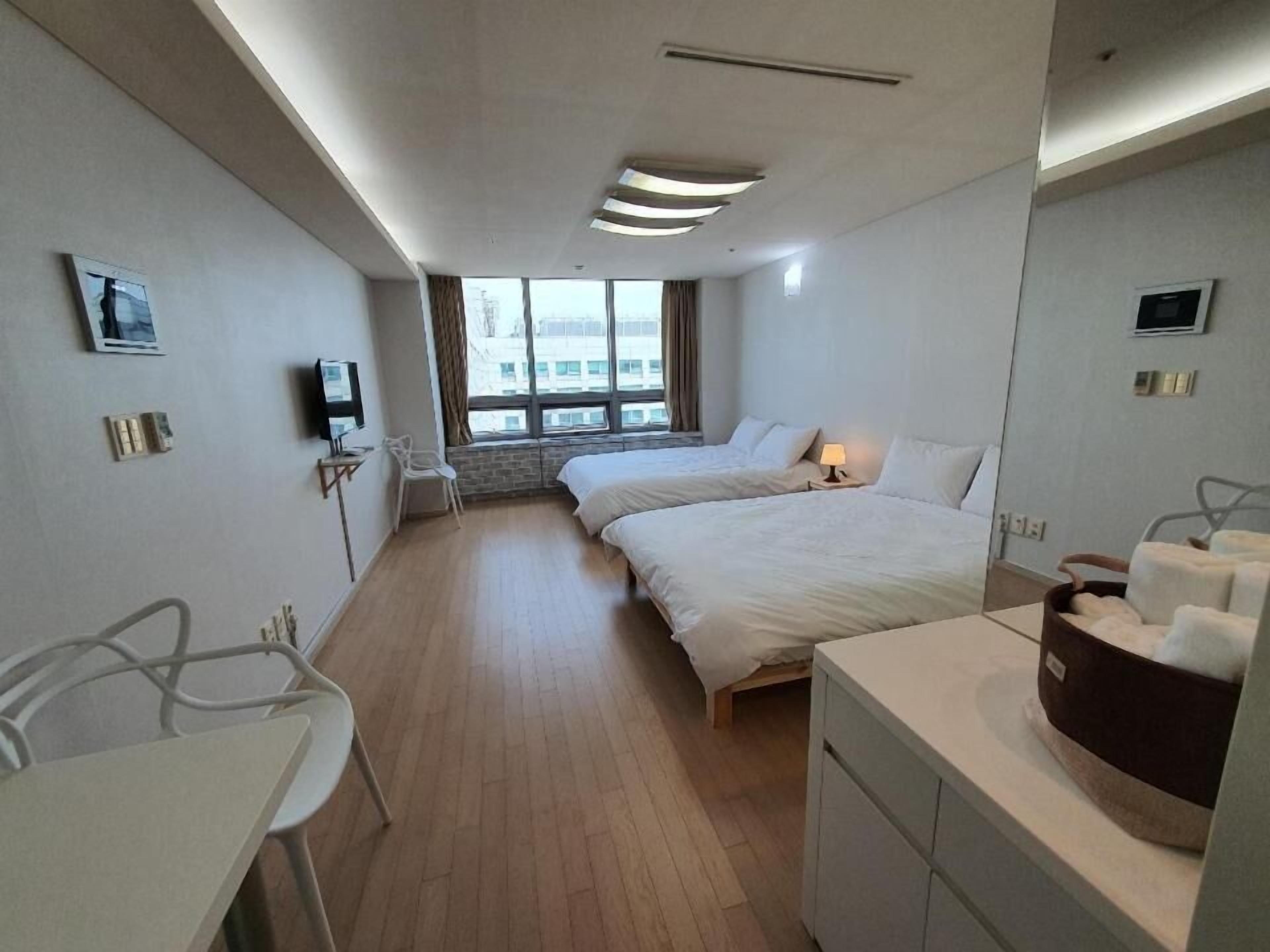 A hotel room with two beds, a desk, a chair, a TV, and a window with a view of buildings.