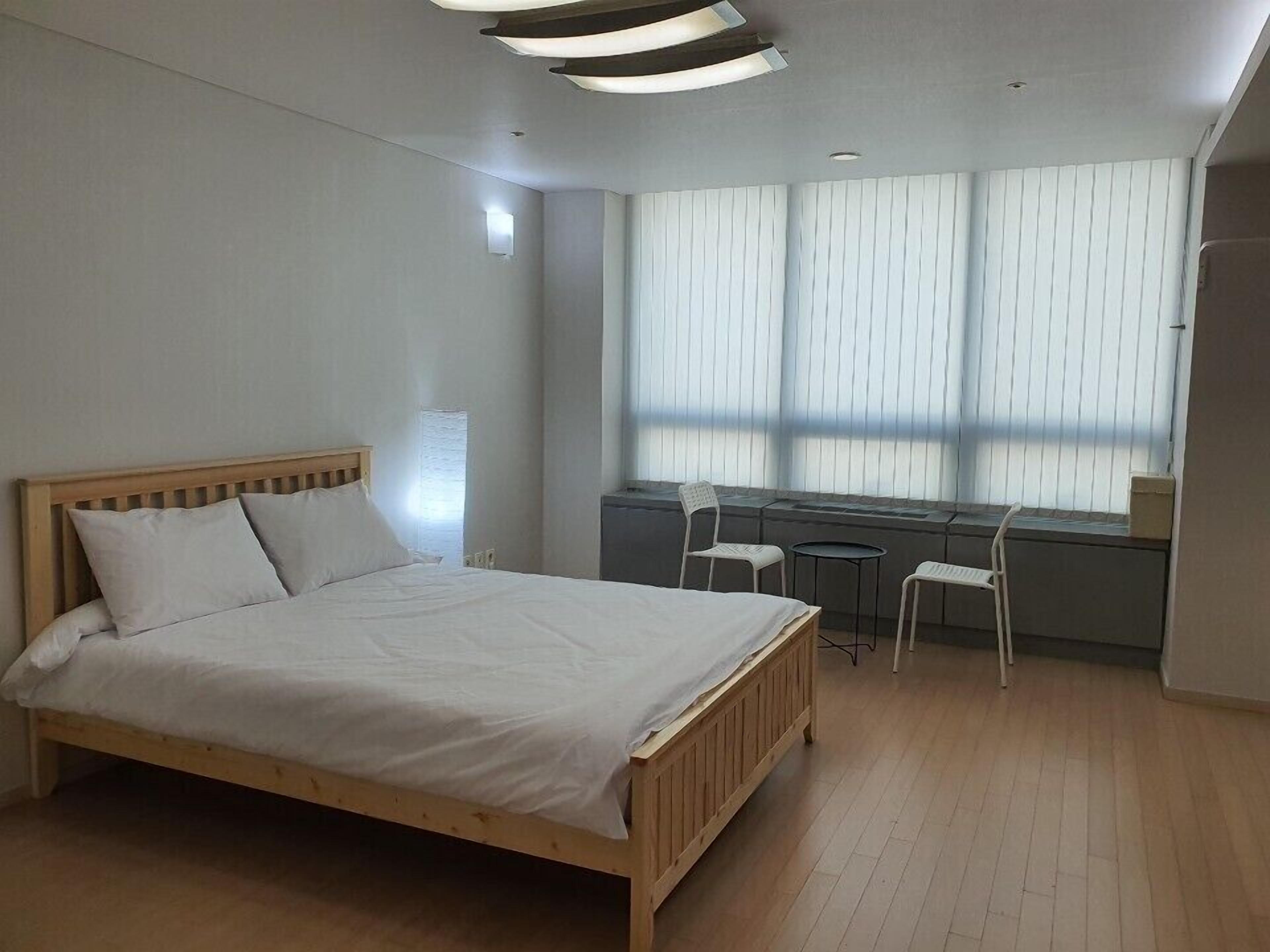 A modern bedroom with a wooden bed, a small desk, two chairs, and a large window with blinds.