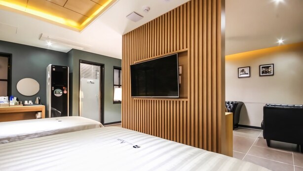 Habitación de hotel con dos camas, un televisor montado en un panel de madera, un escritorio con espejo y un sofá de cuero negro.