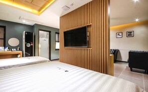 A hotel room with two beds, a TV mounted on a wooden panel, a desk with a mirror, and a black leather couch.