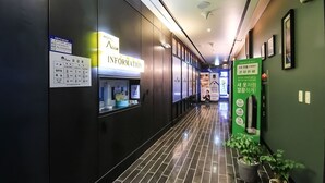 Un pasillo de hotel con mostrador de recepción, tablón de información y una pared verde con un cartel.