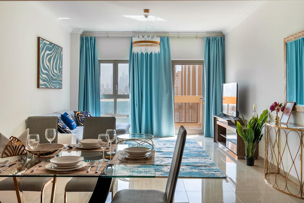 A modern living room with a dining area, large windows with blue curtains, a TV, and a balcony.