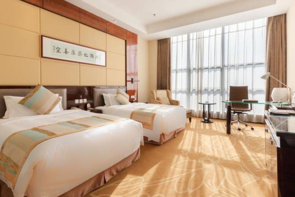 Ein Hotelzimmer mit zwei Betten, einem Schreibtisch, einem Stuhl und einem großen Fenster mit Vorhängen.