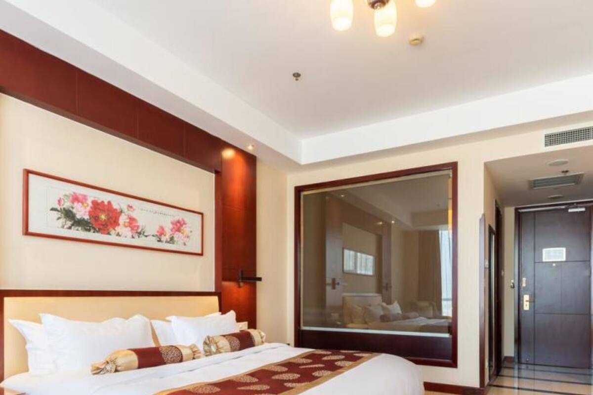 Ein Hotelzimmer mit einem großen Bett, einem Nachttisch, einer gerahmten Blumenarbeit an der Wand und einem großen Spiegel.
