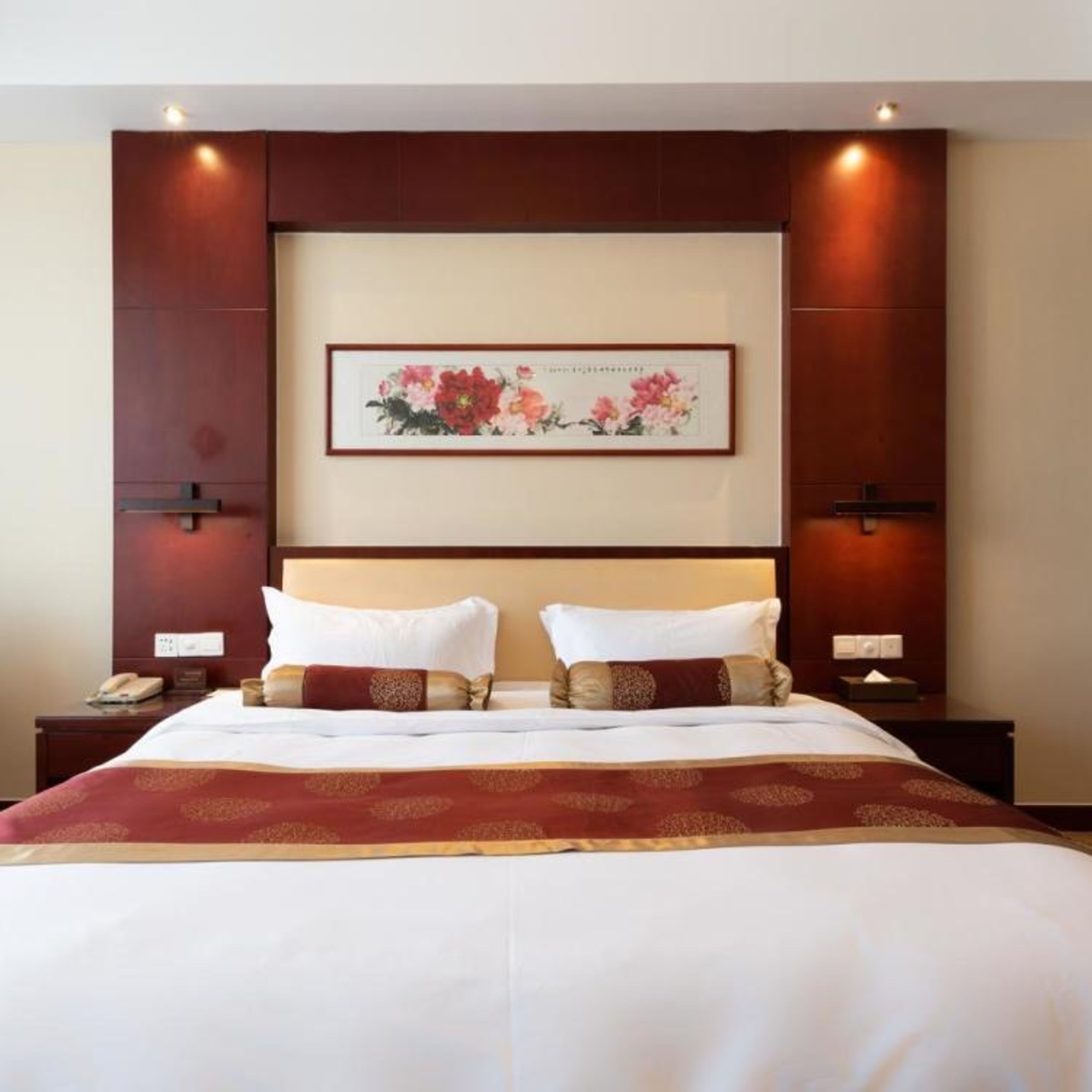 A hotel room with a large bed, two bedside tables, a nightstand, a framed floral artwork on the wall, and recessed ceiling lighting.