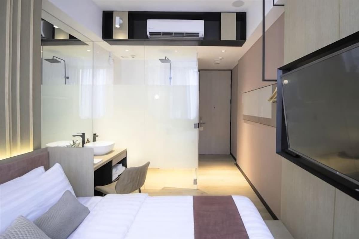 Une chambre d’hôtel moderne équipée d’un lit, d’un grand miroir, d’un téléviseur intégré, d’une salle de bain avec douche et d’un petit coin salon.