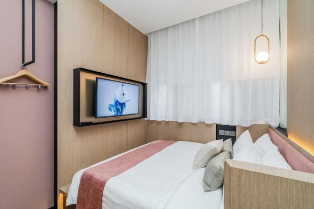 A hotel room with a bed, a television mounted on the wall, a hanging light fixture, and a window with curtains.