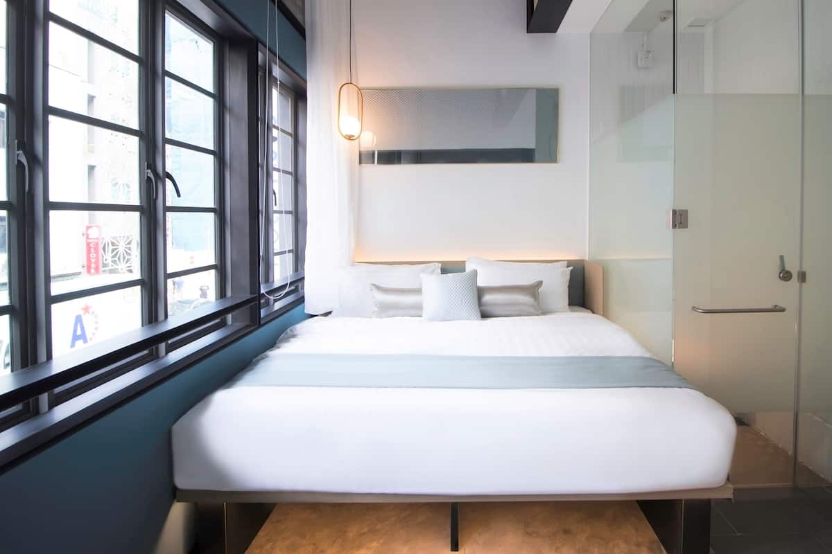 Une chambre d’hôtel moderne, dotée d’un grand lit, d’une table de chevet, d’un miroir et d’une fenêtre donnant sur l’extérieur.
