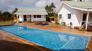 A swimming pool with wooden deck and lounge chairs, surrounded by white buildings with green roofs. - Charlie's Villas (Rarotonga)