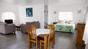 A modern, well-lit living room with a sofa set, a dining table with chairs, a flat-screen TV, and a bed with floral bedding. - Charlie's Villas (Rarotonga)