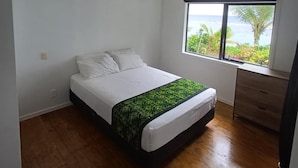Un dormitorio con cama, ventilador de techo, ventana con vista al océano y cómoda de madera.