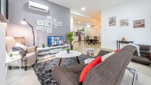 A modern living room with a grey sofa, a brown sofa, a white coffee table, and a TV on a white stand. There are framed pictures on the wall and a potted plant on the coffee table. - Comfort & Cozy Family Suite (Ampang)