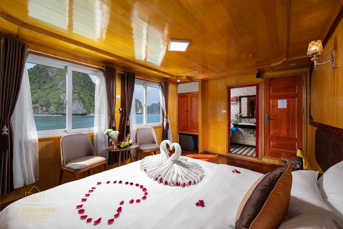 A luxurious cabin with a bed adorned with a swan-shaped arrangement of rose petals, a view of the sea, and a wooden interior.