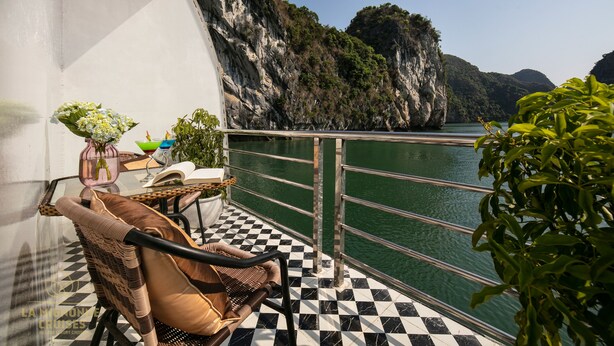 A balcony with a checkered floor, wicker furniture, and a view of a body of water and cliffs. - La Mignonne Cruises (Tuan Chau)