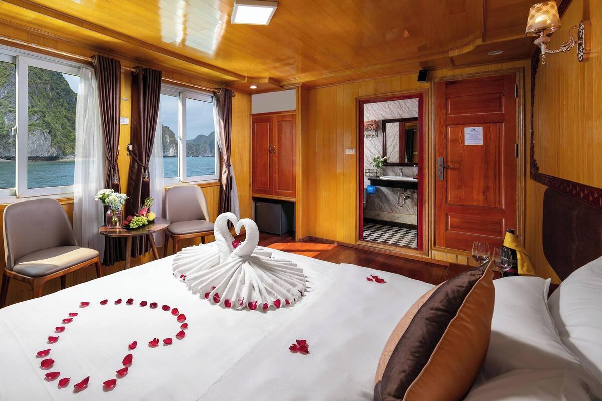 A room with a bed, a table with a vase of flowers, a chair, a view of the sea, and a bathroom door.