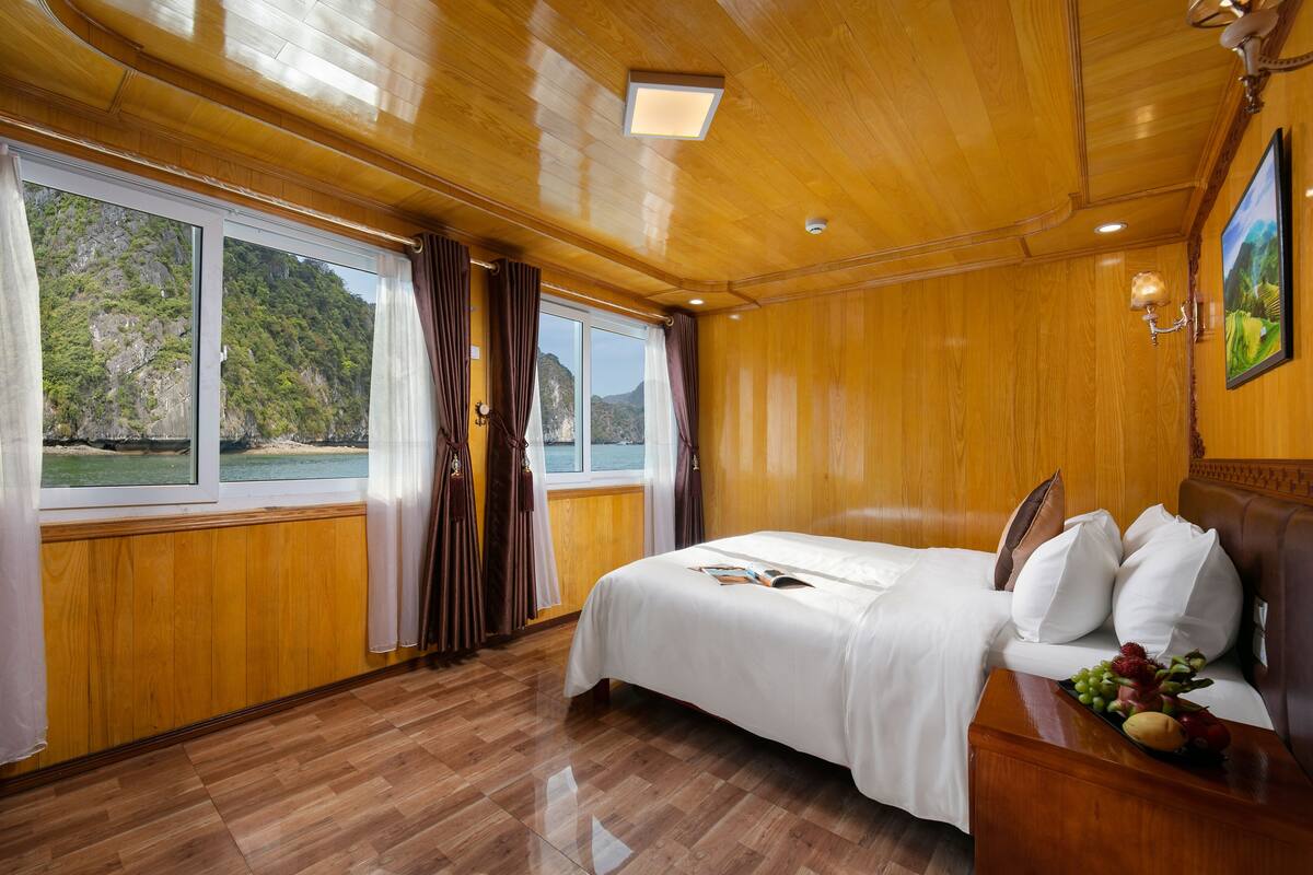 A wooden-paneled room with a bed, a nightstand with fruit, a window with curtains, and a painting on the wall.