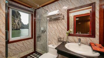 Un baño con pared de mármol, ventana con vista a un acantilado, inodoro blanco, lavabo y espejo.